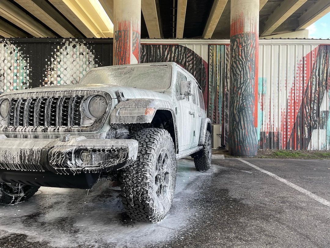 A white Jeep covered in thick car wash foam parked under a concrete overpass with painted pillars.