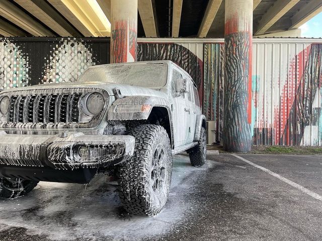 White Jeep being washed under an overpass, covered in soap foam, near colorful pillars