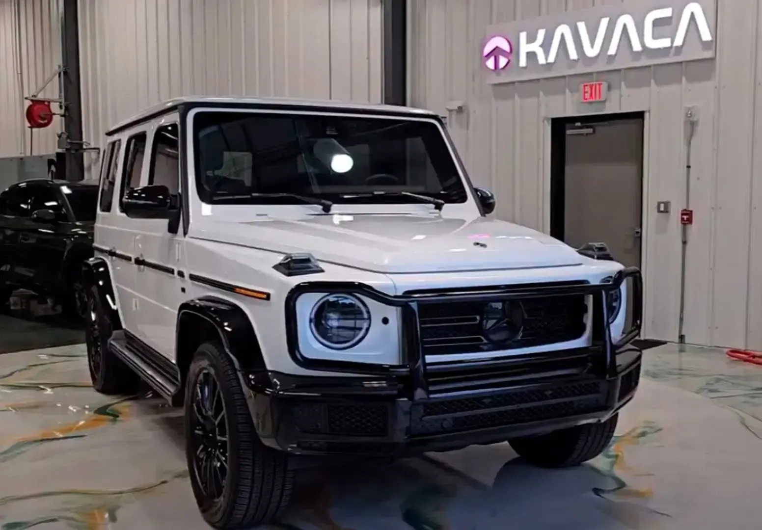 White Mercedes-Benz G-Class SUV in a garage, front view, with black trim and a Kavaca sign in the background