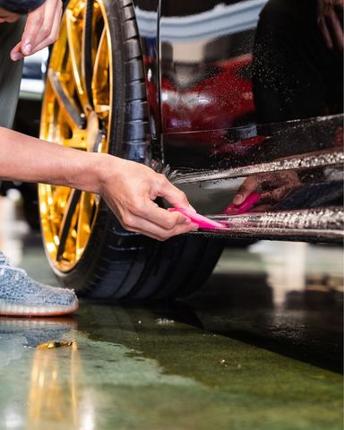 Hand washing a black car’s side skirt with a pink brush next to a gold wheel in a wet garage