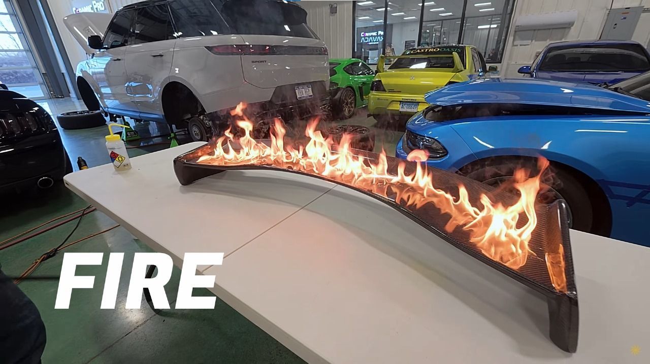 A curved metal piece burns with open flames on a white table in a garage surrounded by various cars.