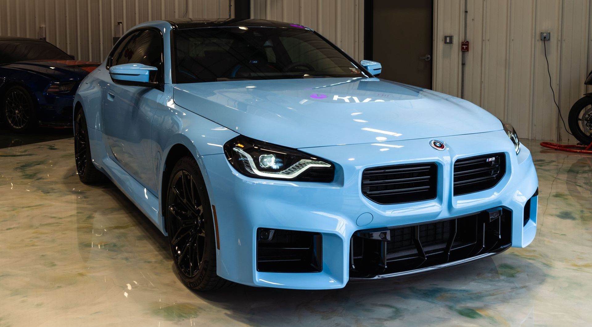 A light blue BMW M2 sports car parked inside a garage with black wheels and a black roof.