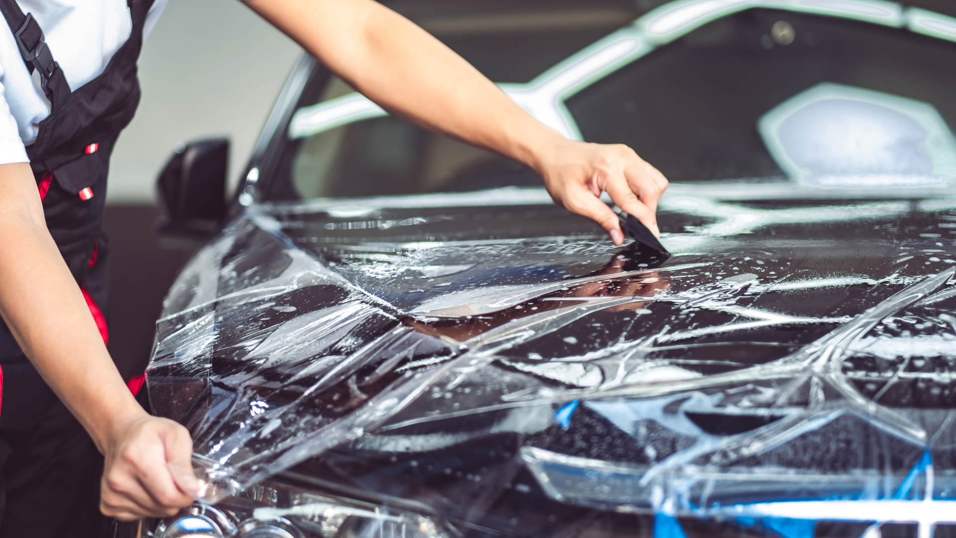 A person in uniform uses a squeegee to apply clear protective film to the black hood of a car.