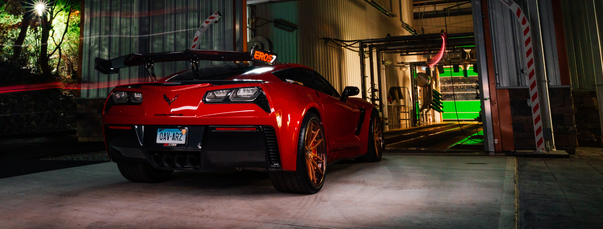 A red Chevrolet Corvette with a large rear spoiler parked at the entrance of a glowing green car wash at night.