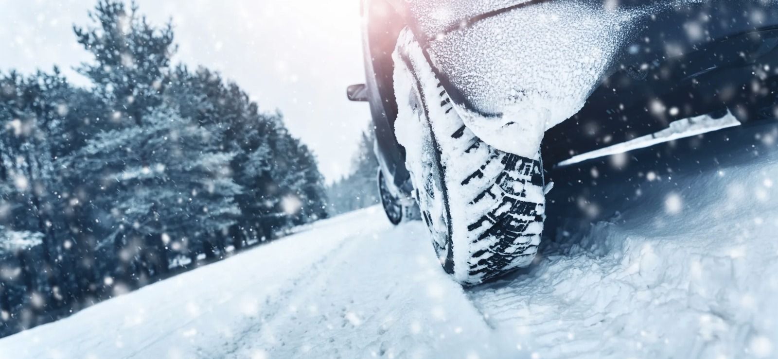 A close-up view of a car tire with a winter tread pattern driving on a snowy road with trees in the background.