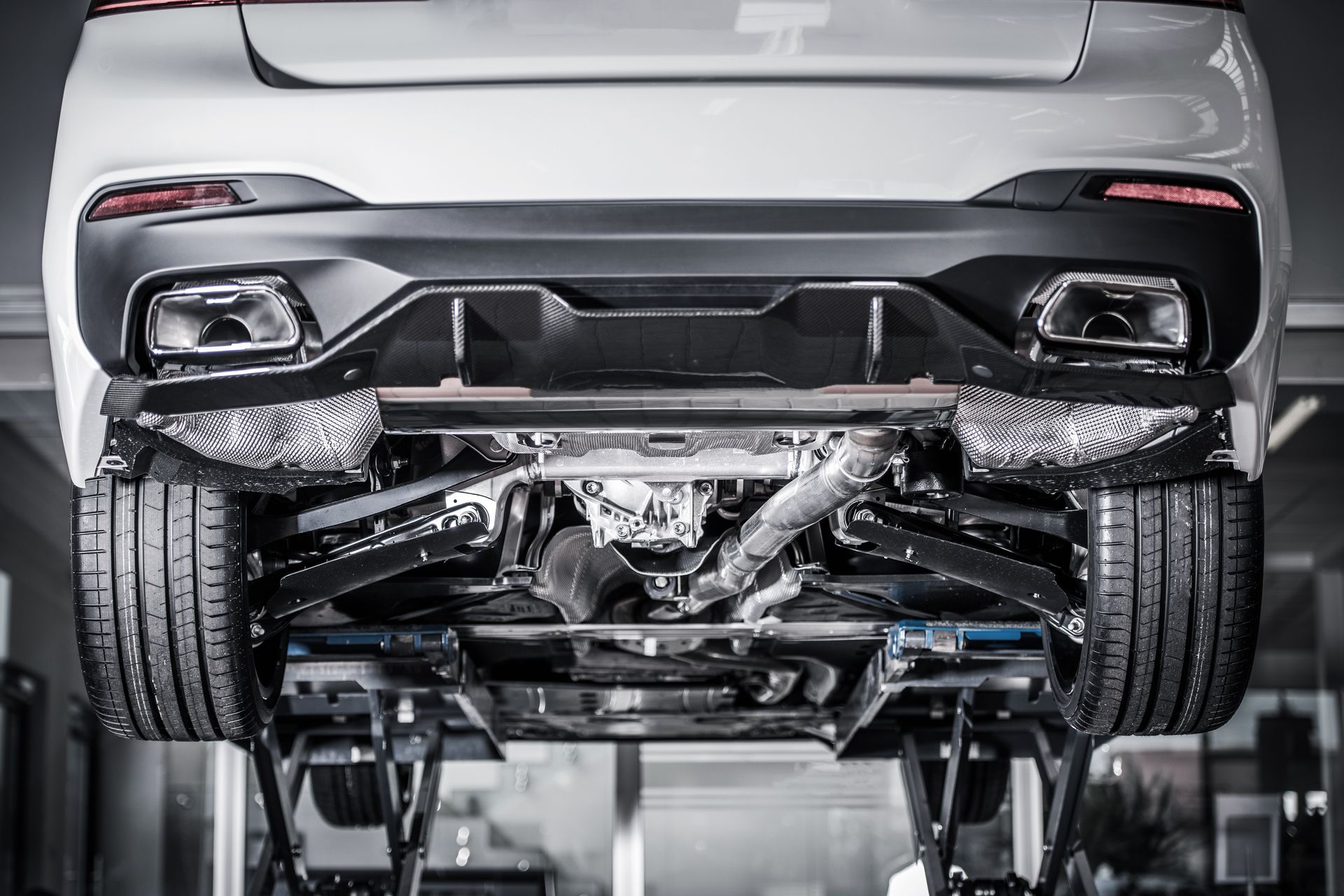 A white car viewed from underneath while elevated on a mechanical lift in a garage, showing its chassis and exhaust.