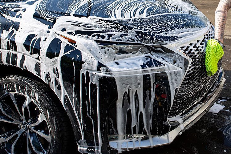 A person uses a bright green sponge to wash a dark-colored Lexus covered in thick white soap suds.