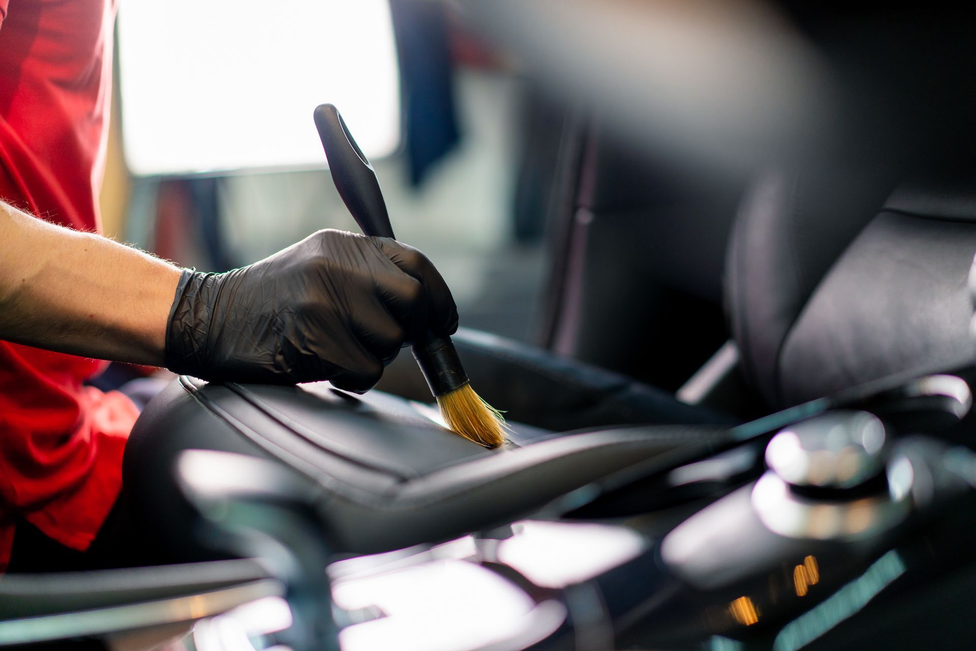 A person in a red shirt and black glove uses a small brush to clean the leather surface of a car interior.