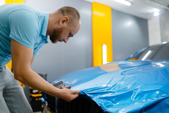 Mechanic applying blue vinyl wrap to a car hood in a garage