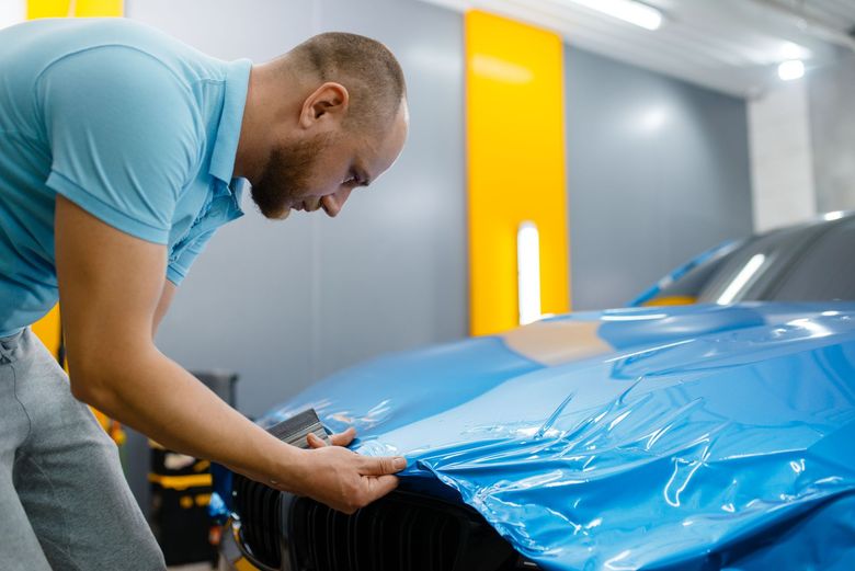 A technician applies a bright blue vinyl wrap to the hood of a car inside a brightly lit workshop.