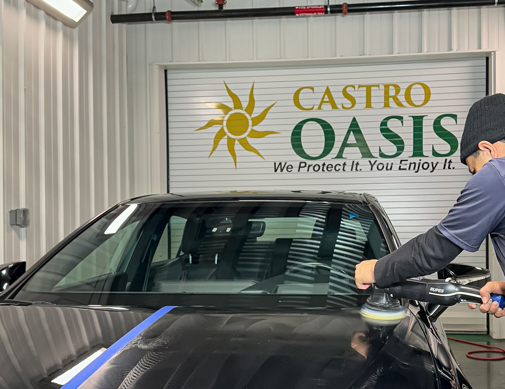 A worker uses a power polisher on the hood of a black car in a garage with a Castro Oasis logo on the back wall.