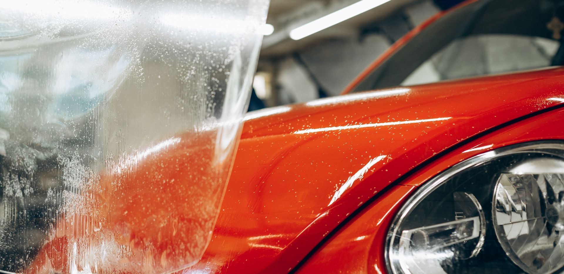 A clear protective film is being applied to the bright red hood of a car in a workshop.