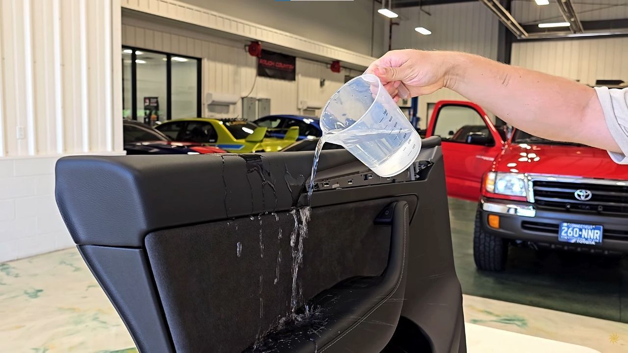 A person pours water from a measuring cup onto a black car door panel in an auto shop to test its water resistance.