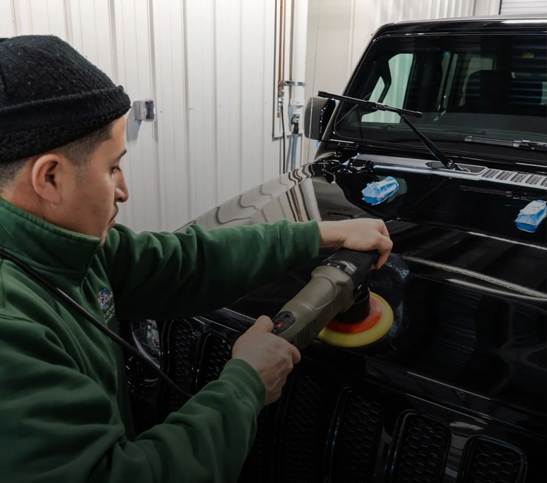 A person in a green sweater uses a power buffer to polish the black hood of a vehicle in an indoor shop.