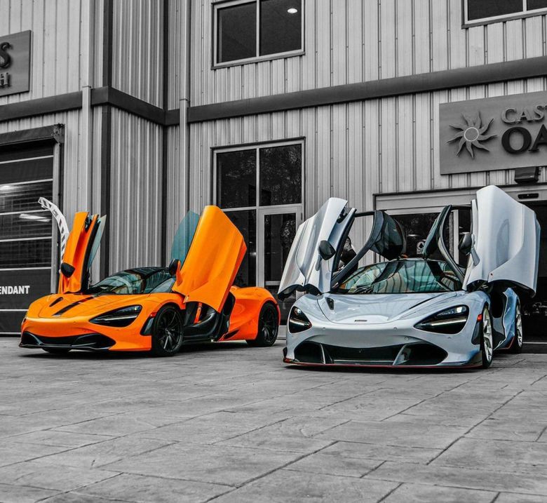 An orange and a light grey McLaren supercar with their doors open, parked in front of a modern industrial building.