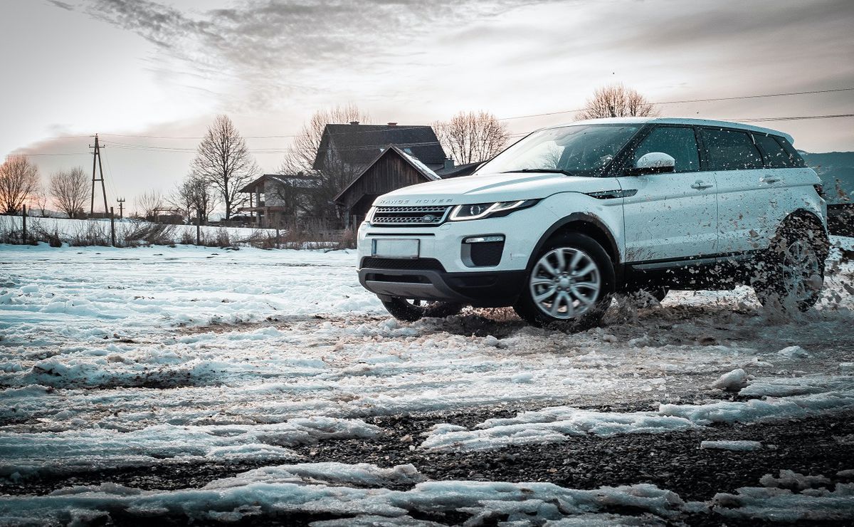 A white Range Rover SUV driving through slush and snow in a rural landscape with buildings in the background.