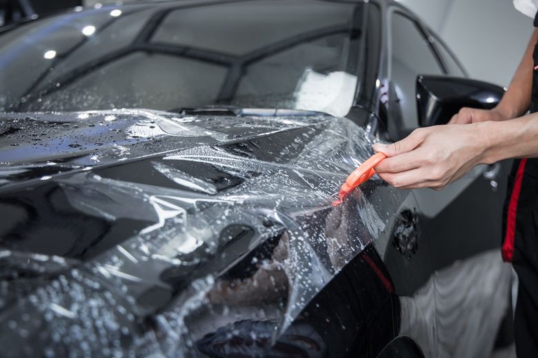 A technician uses a tool to apply clear protective film to the dark hood of a vehicle.