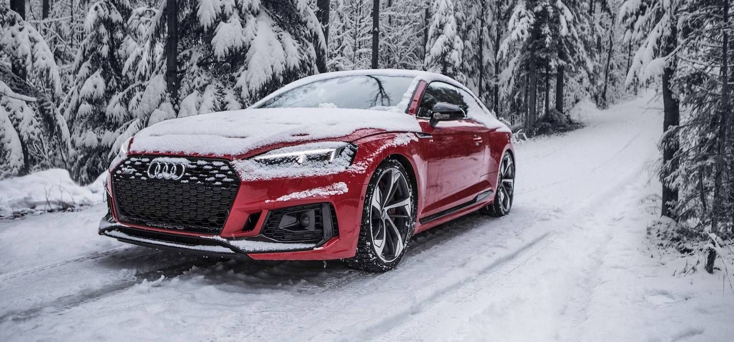 A red Audi coupe parked on a snowy road surrounded by a dense, snow-covered forest.