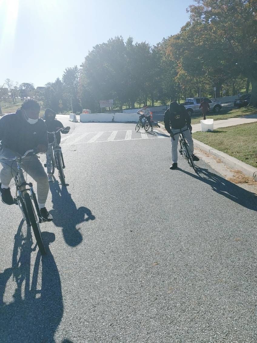 JCB Academy Bike Club - Principals on bicycles on a paved road. Trees and a building are in the background. Sunny day.