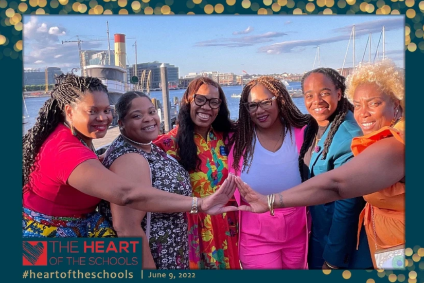 Heart of School Honors - Six women outdoors, smiling, with hands forming a triangle. Buildings and water in background.