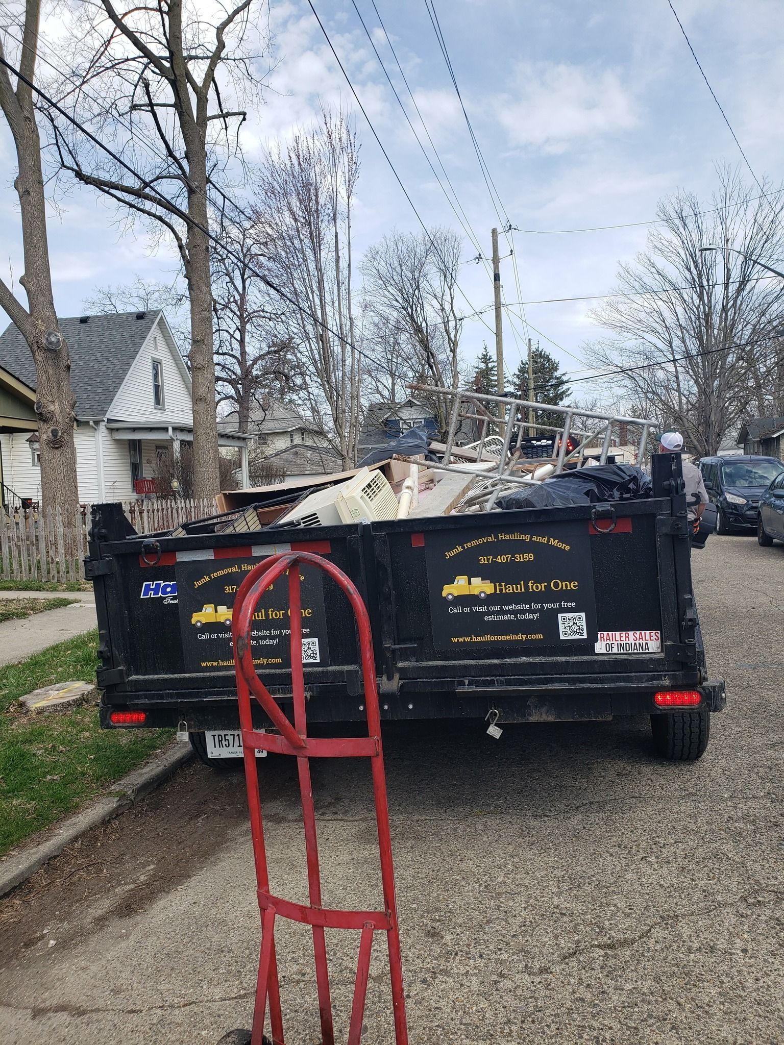 A dumpster is parked on the side of the road next to a red dolly. A dumpster is parked on the side of the road next to a red dolly.