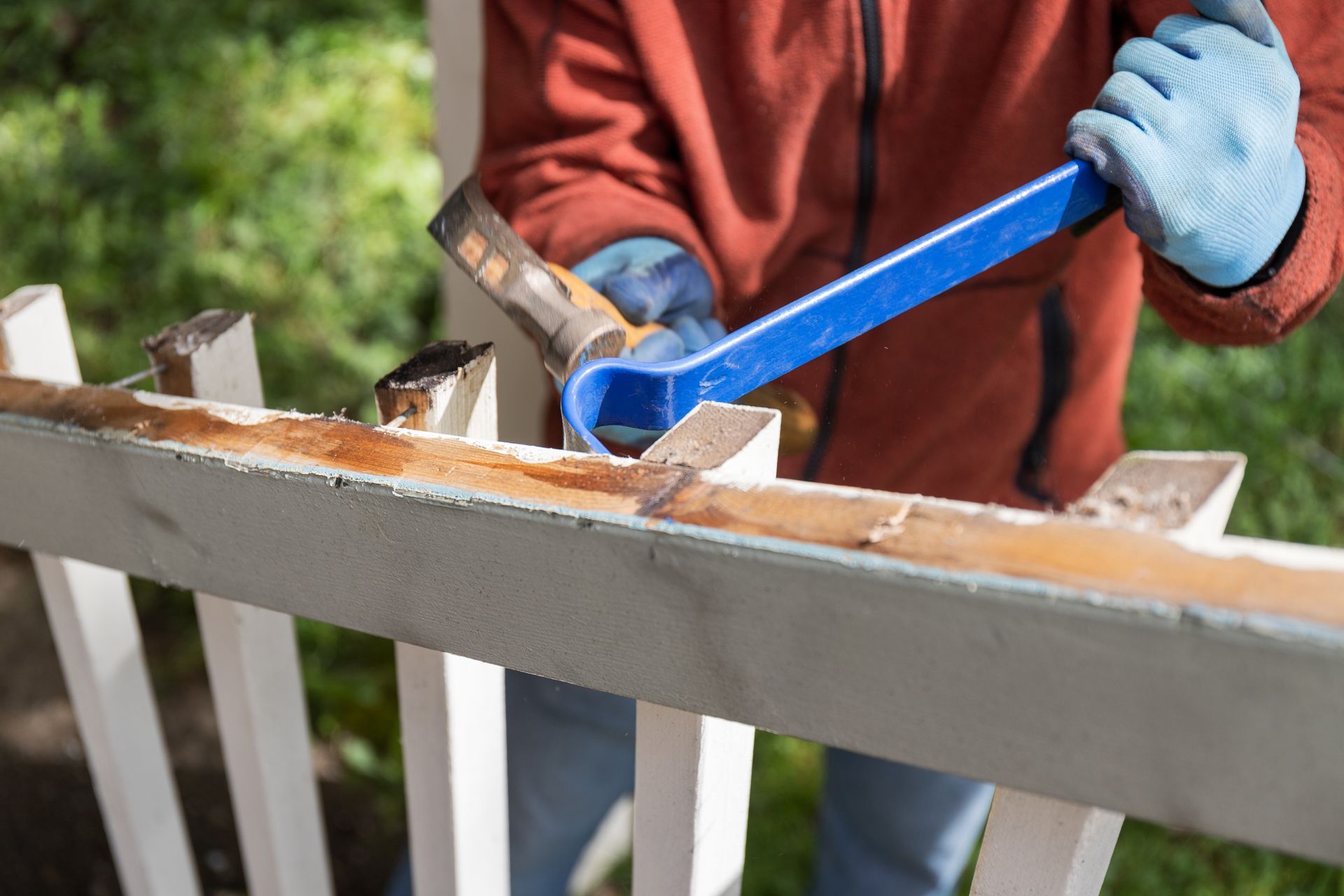 A man is using a pry bar to remove rust from a white picket fence.