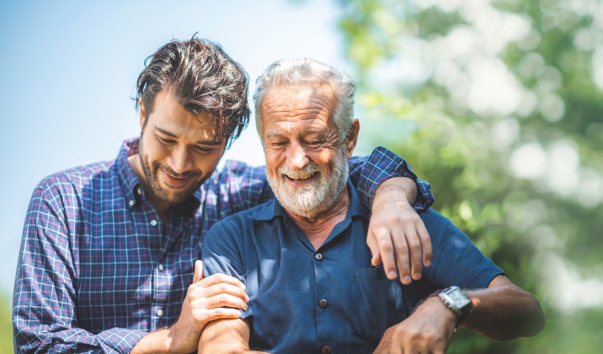 A younger man with his arm around an older man, smiling. They are outdoors in a green, blurred background.