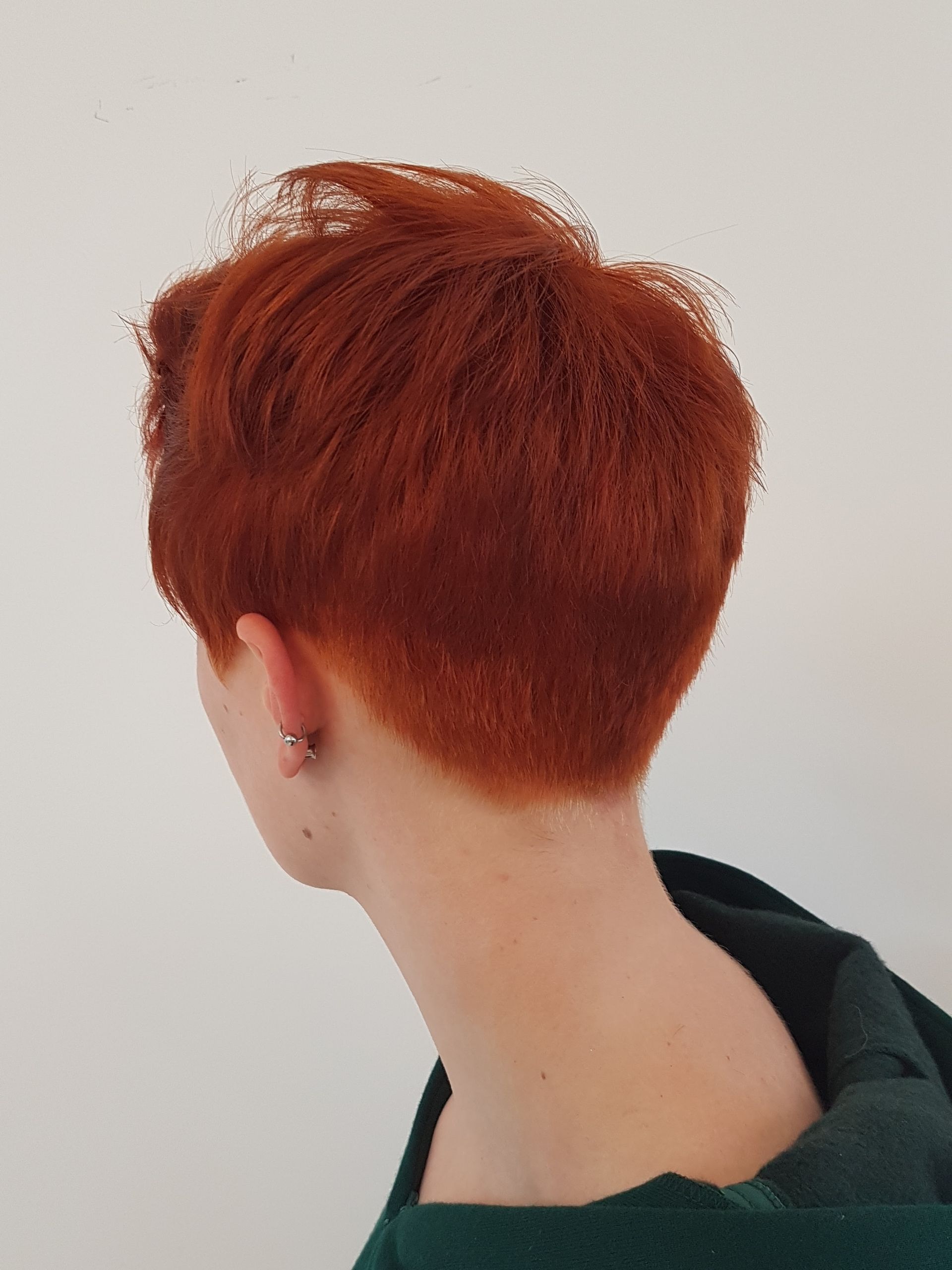 Achteraanzicht van een persoon met kort rood haar en een oorbring, gekleed in een donkere top, tegen een witte muur.