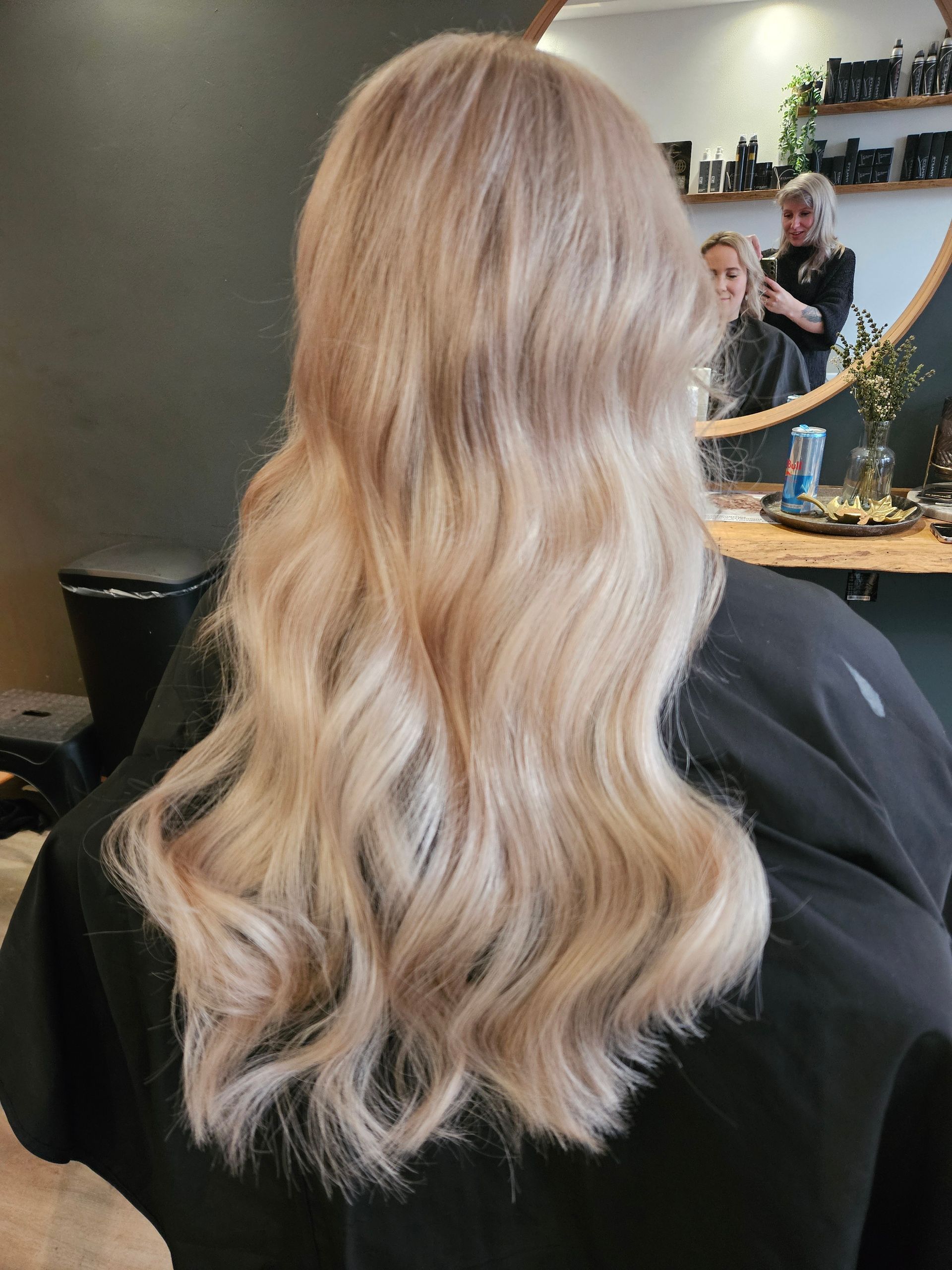 Blond, golvend haar, van achteren gezien in een kappersstoel, met een spiegel en de stylist op de achtergrond.
