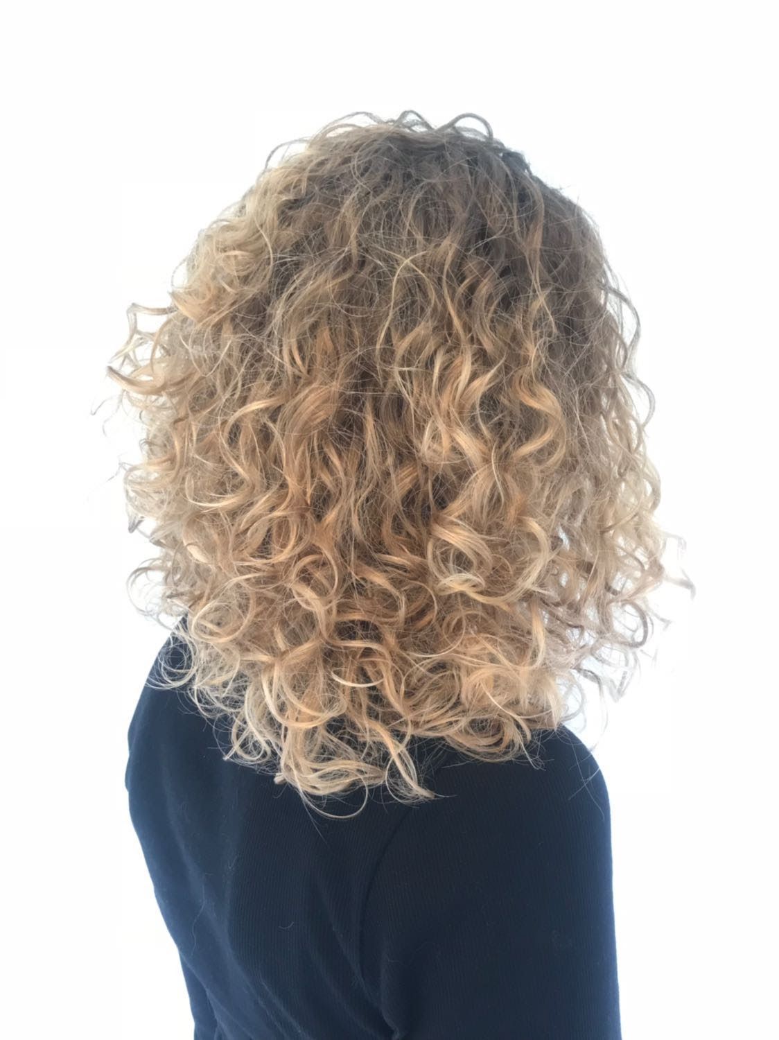 Achteraanzicht van een persoon met krullend blond haar, gekleed in een donkere top, tegen een witte achtergrond.
