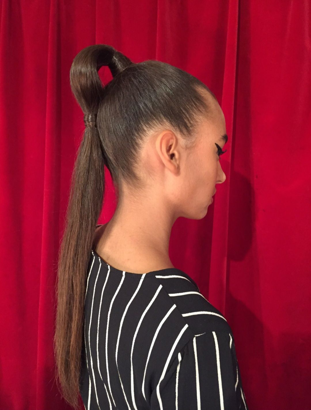 Zijprofiel van een vrouw met een lange, hoge paardenstaart tegen een rode gordijnachtergrond, gekleed in een zwart gestreept topje.