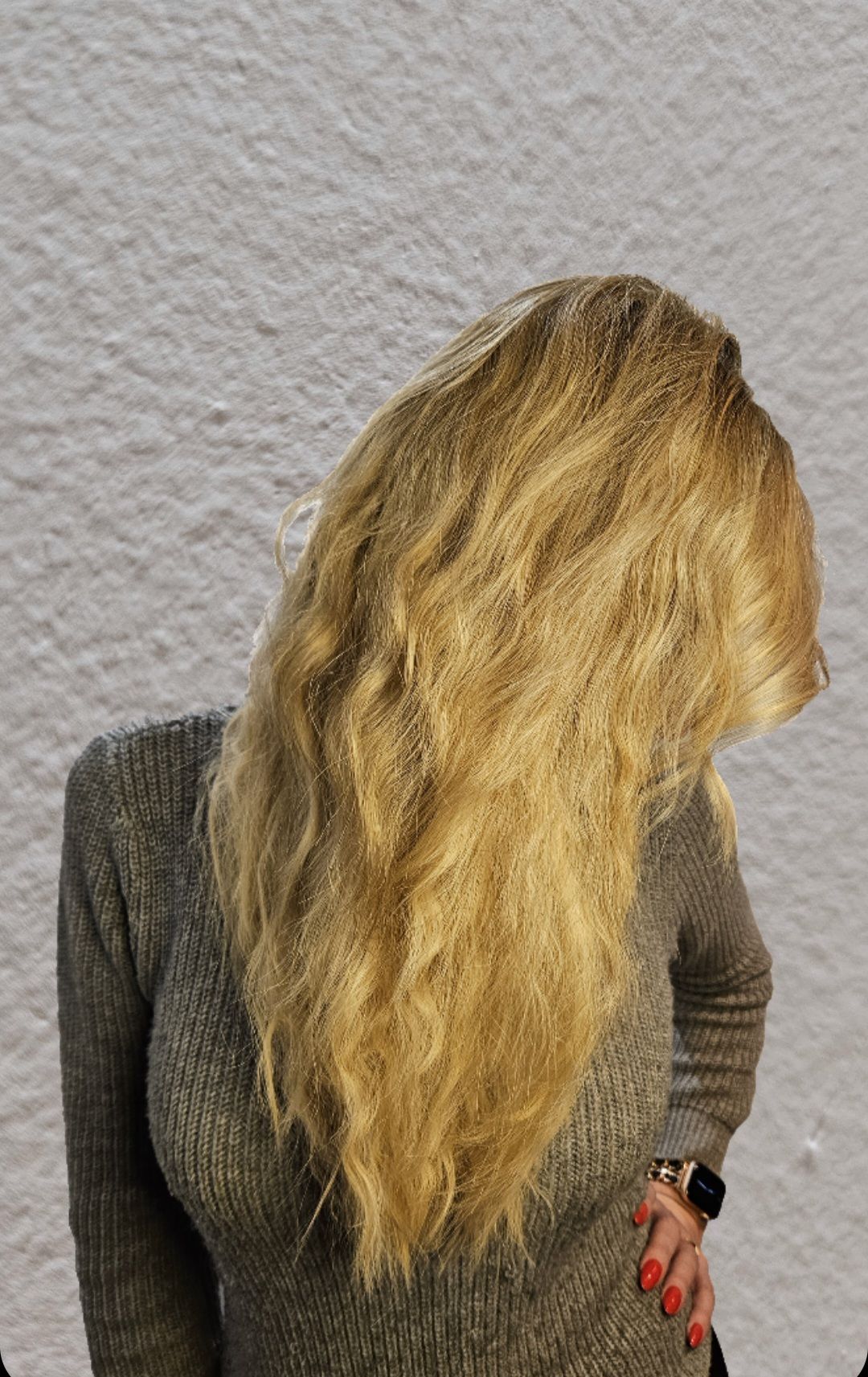 Een blondharige persoon in een grijze trui staat zijdelings tegen een lichte, gestructureerde muur.