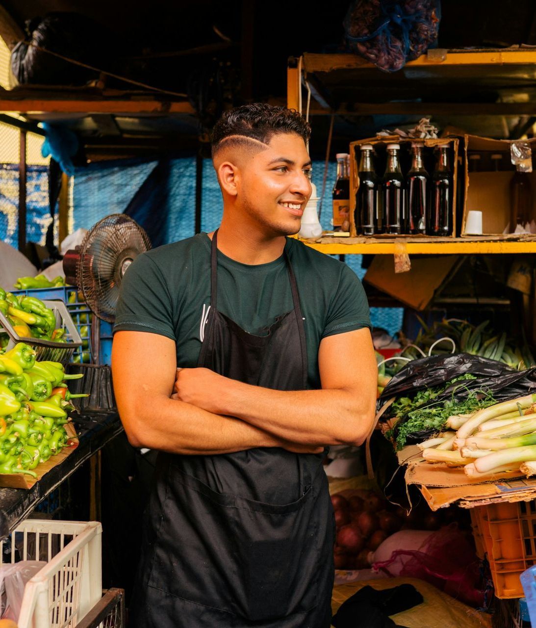 A man in an apron is standing in front of a vegetable stand.