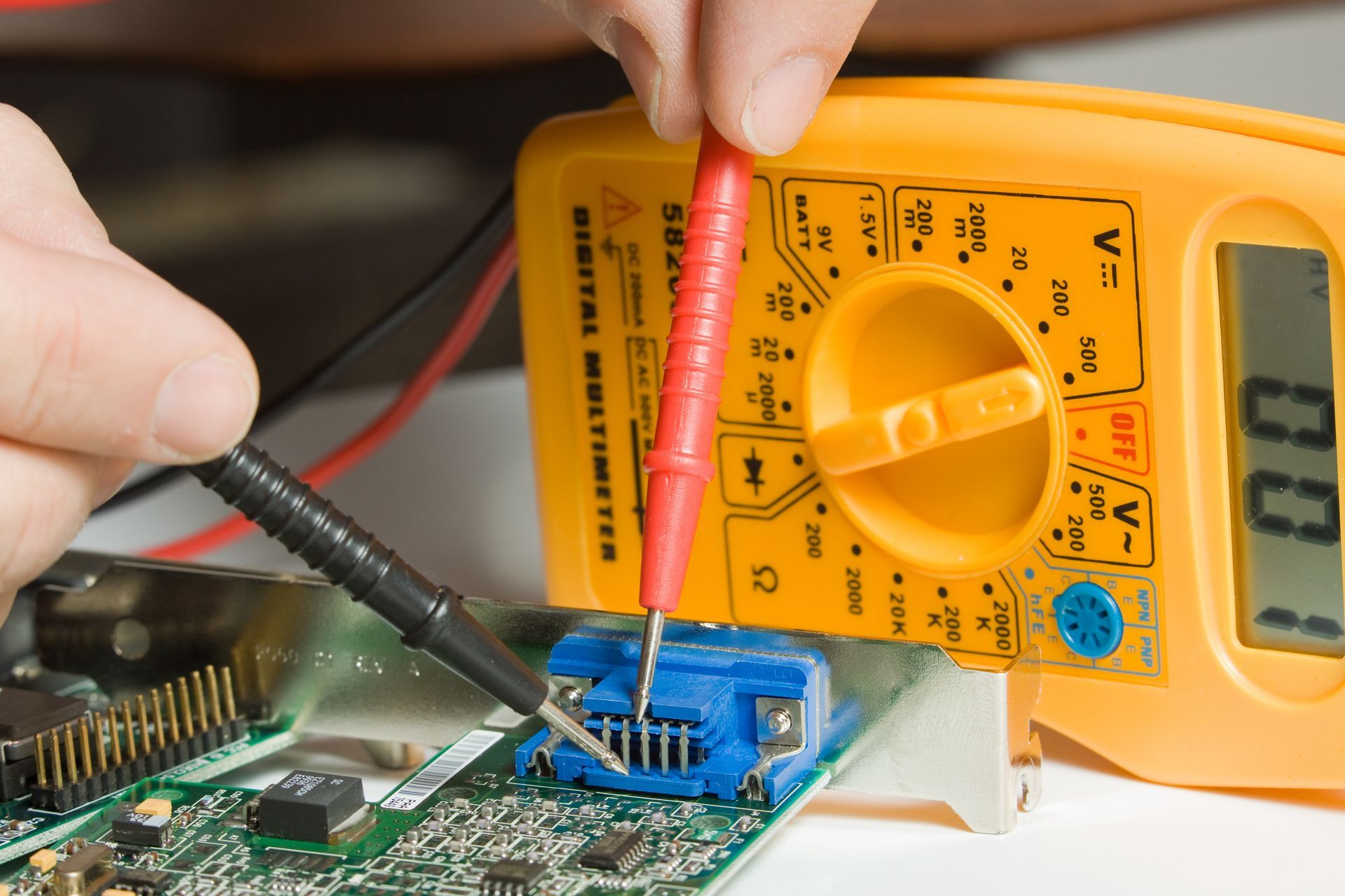 Hands testing a circuit board with red and black multimeter probes beside a yellow digital meter