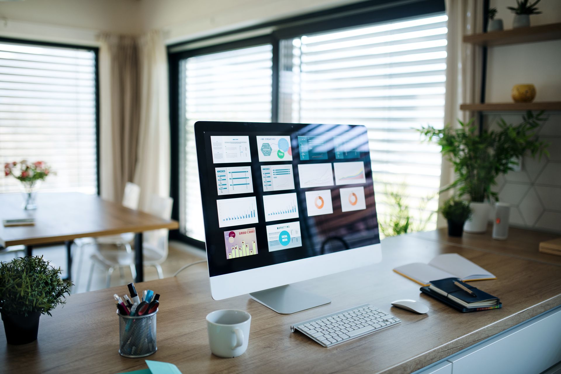 Modern home office desk with iMac, keyboard, notebooks, coffee mug, and plants by bright windows