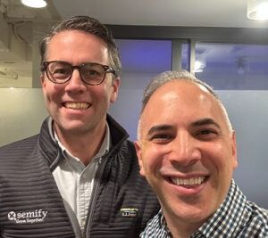 Two smiling men posing indoors, one in a Semify jacket and one in a checkered shirt.