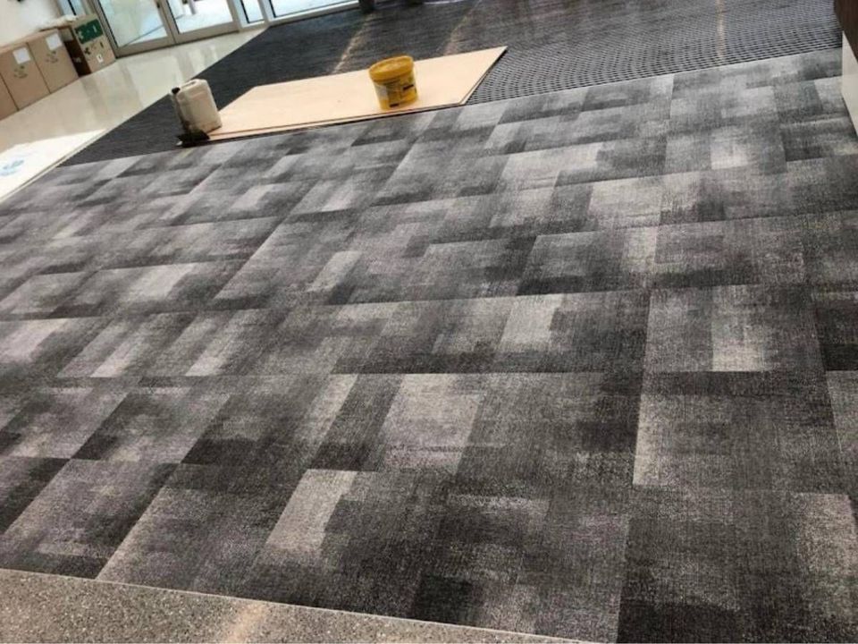 Grey tonal commercial carpet tiles part-laid across a floor with a roller and adhesive alongside