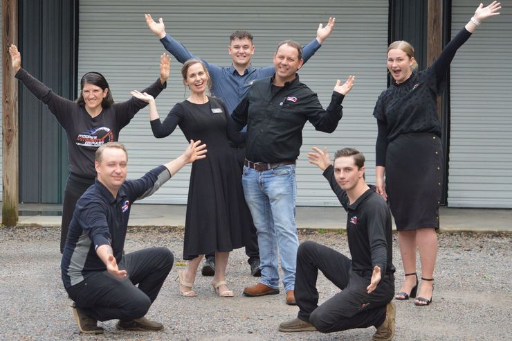 Seven people in black outfits posing outdoors on steps with arms raised, smiling.
