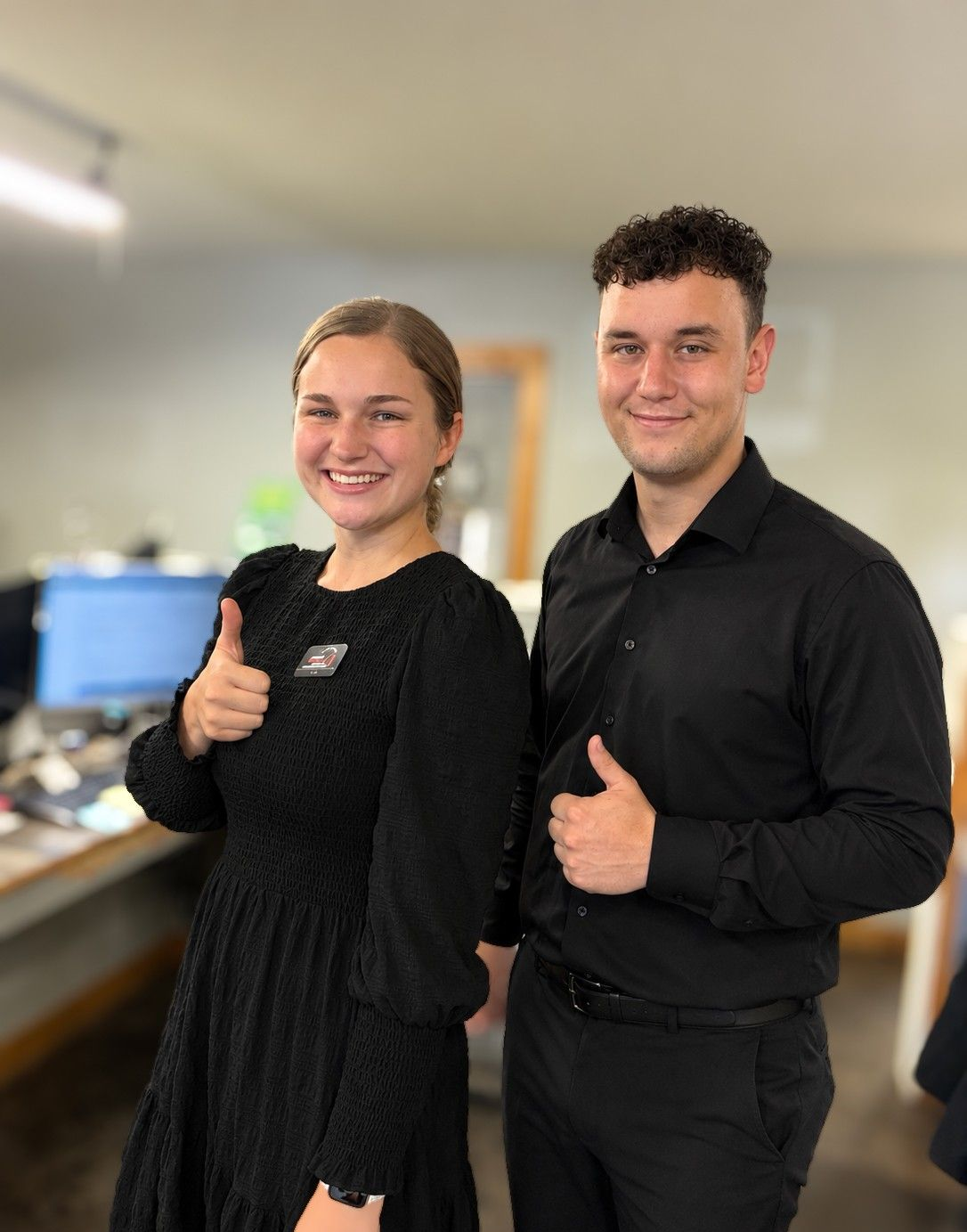 Two smiling coworkers in black outfits giving thumbs-up in an office setting