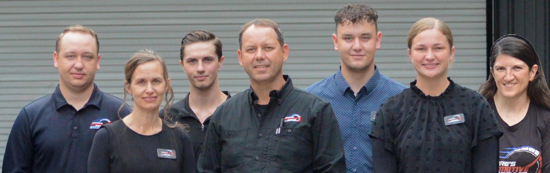 Seven people in matching dark shirts standing together in front of a gray wall.