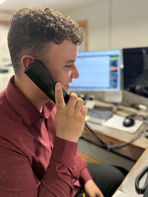 Man in a red shirt speaking on a phone at a desk with computer monitors in the background