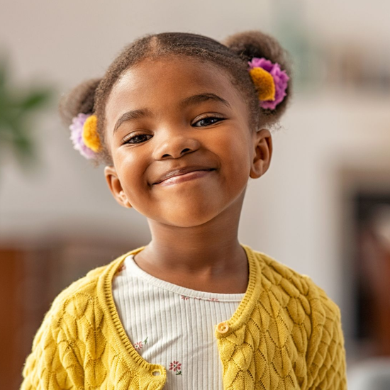 Smiling child wearing a yellow cardigan with flower hair ties, standing indoors