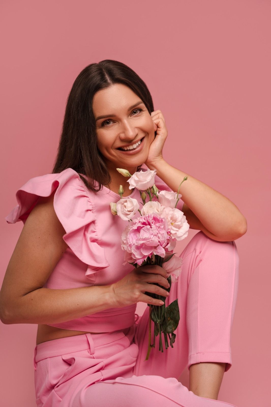 Mujer sonríe, sosteniendo flores rosas, vistiendo una blusa y pantalones rosas, sobre un fondo rosa.