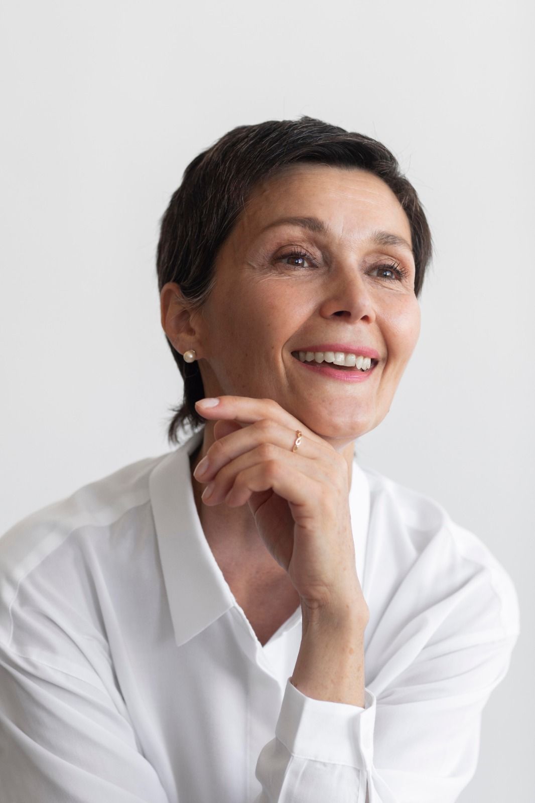 Mujer sonriendo, con la mano en la barbilla, vestida con camisa blanca, mirando hacia arriba.