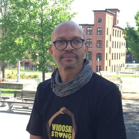 Person wearing glasses and a black shirt stands outdoors in a park with trees and brick buildings.