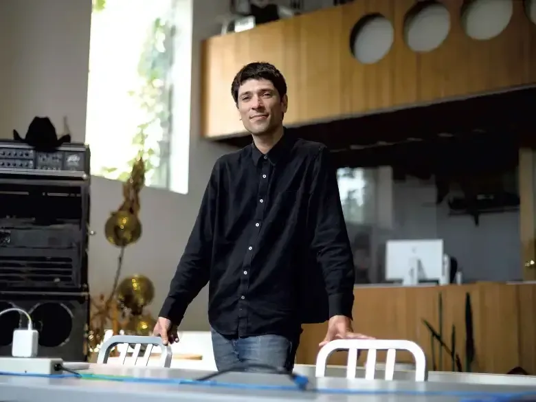 Man standing in a modern room beside a table and chairs, with audio equipment and wood-paneled walls behind him