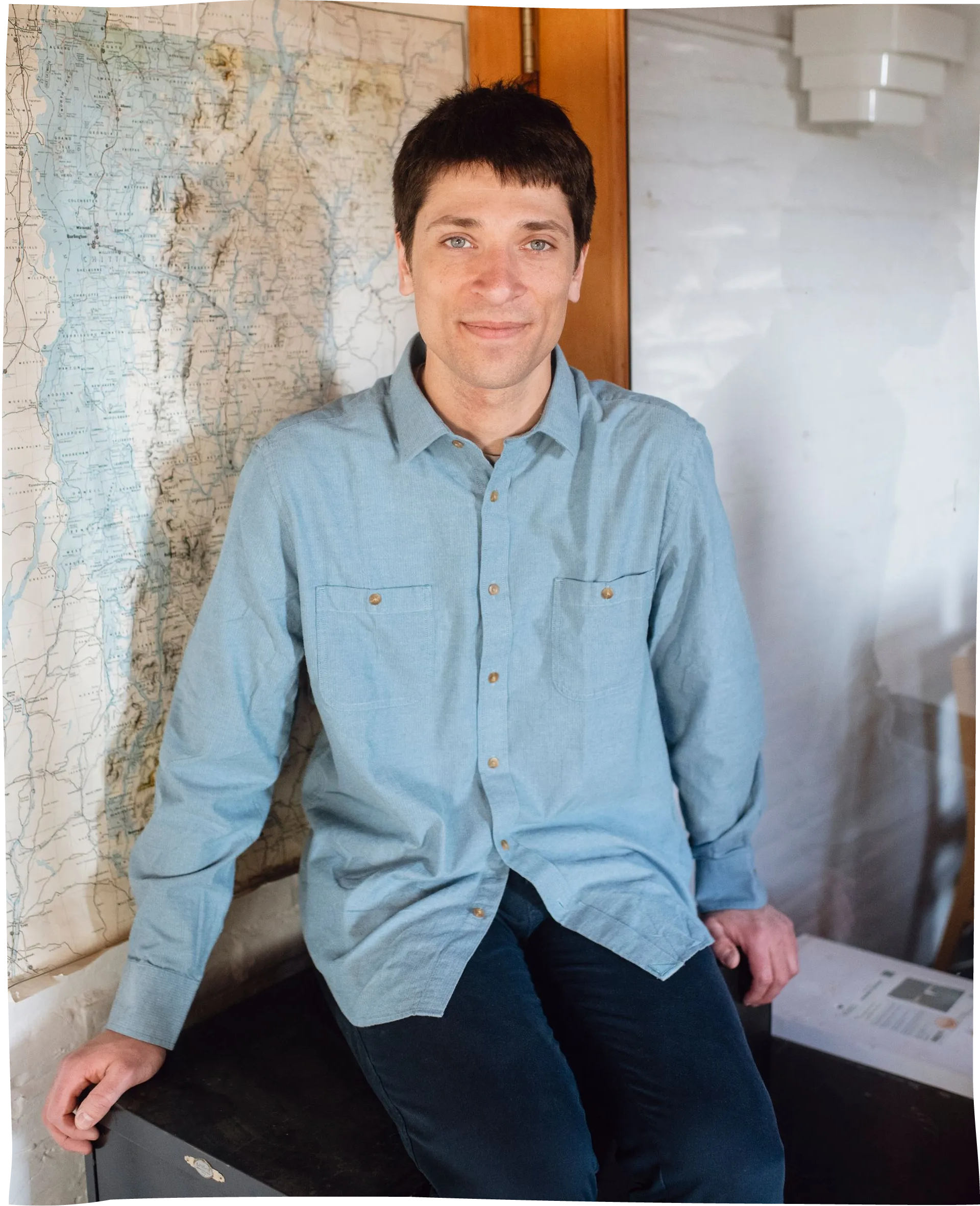 Man seated indoors beside a wall map, wearing a light blue shirt and dark pants, near a wooden doorway