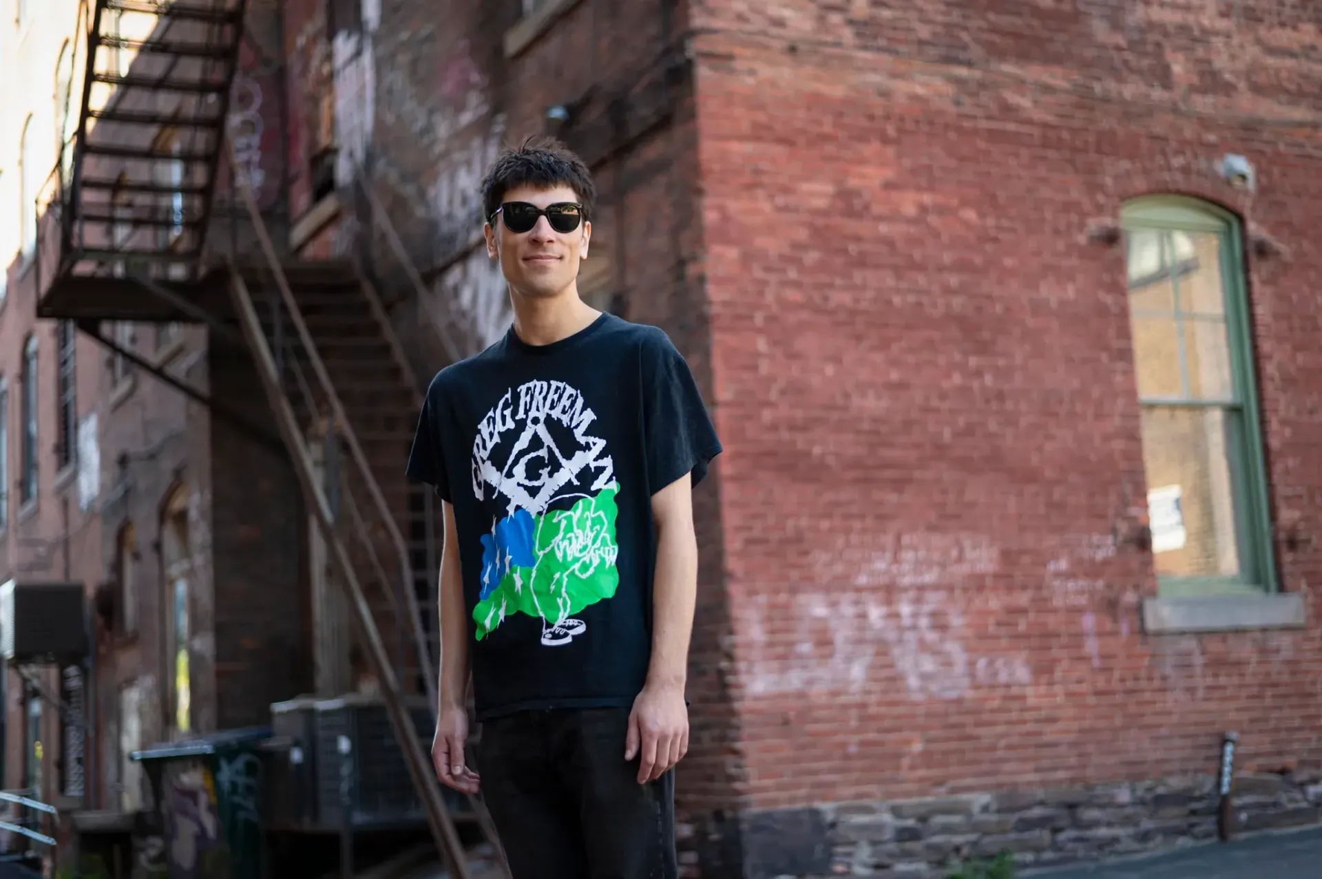 Person in sunglasses stands by a brick wall and fire escape, wearing a black graphic T-shirt.