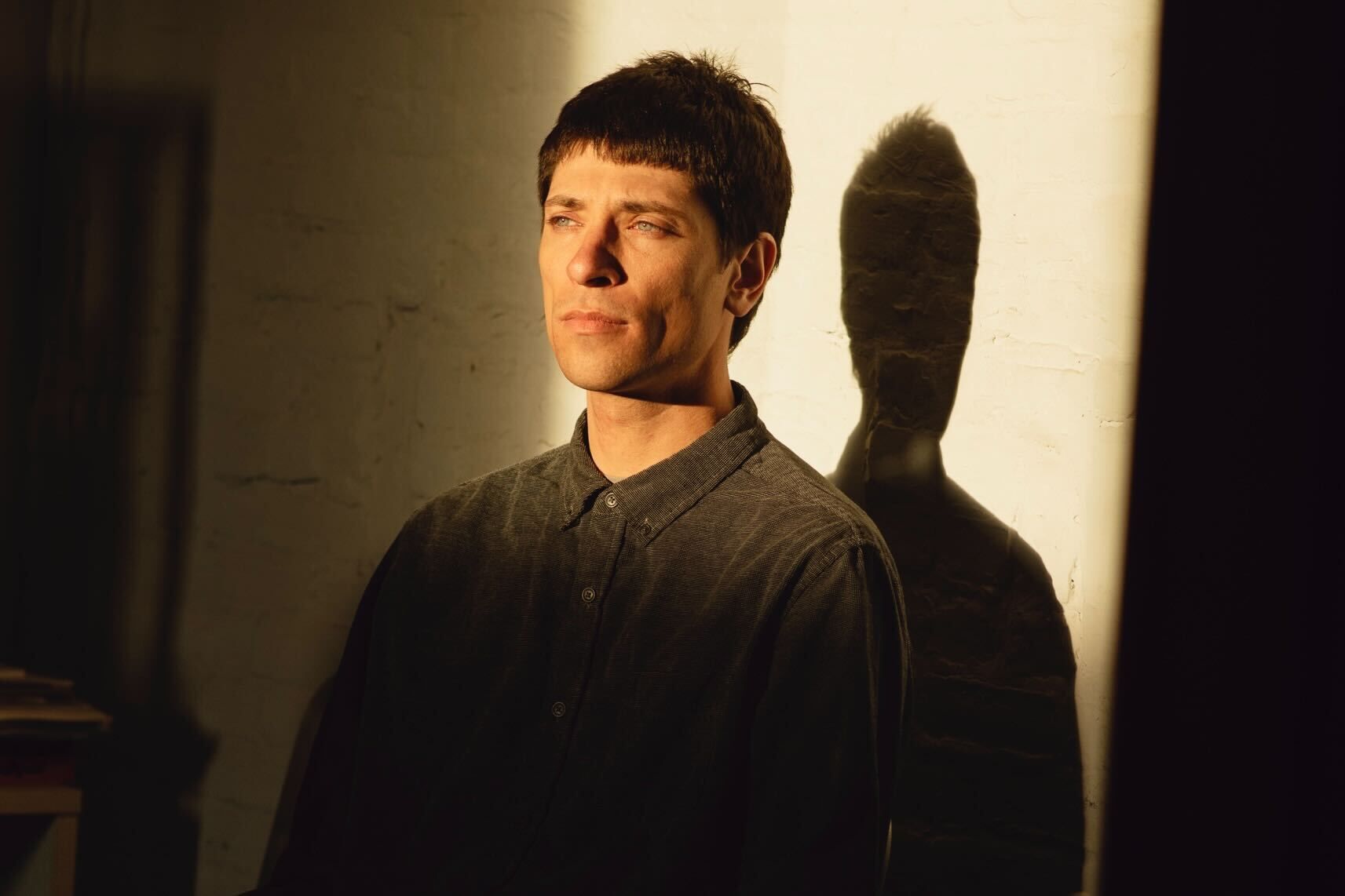 Man standing against a sunlit wall, casting a sharp shadow, with a serious expression.