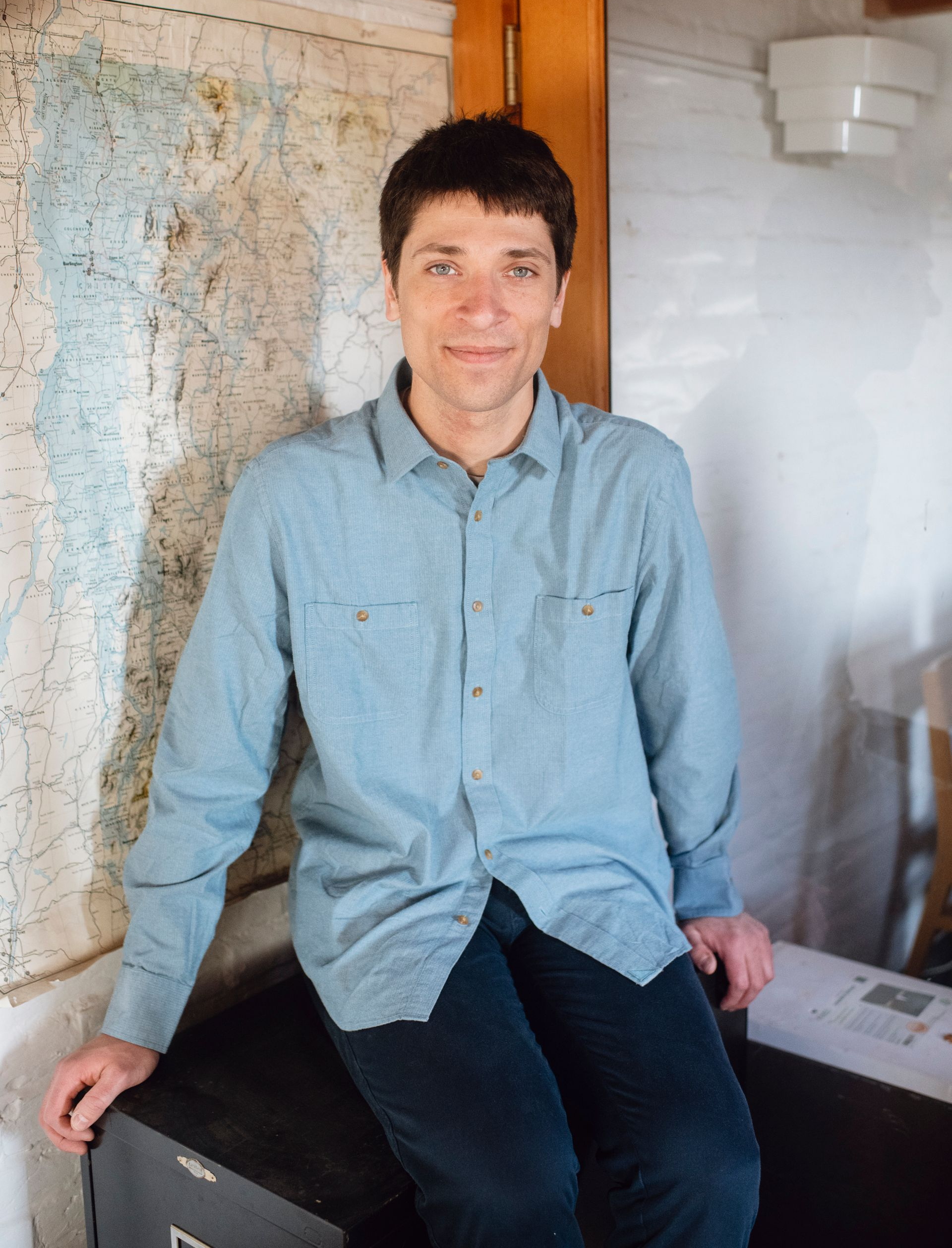 Man seated indoors beside a wall map, wearing a light blue shirt and dark pants, near a wooden doorway